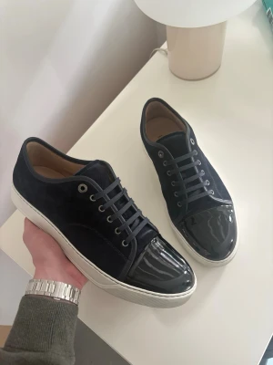 Mörkblå Lanvin skor - Snygga mörkblåa Lanvin patent skor.  Skorna har vita sulor, mörkblå skosnören och silverfärgade öljetter. Insidan är beige och designen är stilren och modern, perfekt för dig som gillar en clean look. Skorna är knappast använda då storleken inte passade mig bra. Storlek 45!