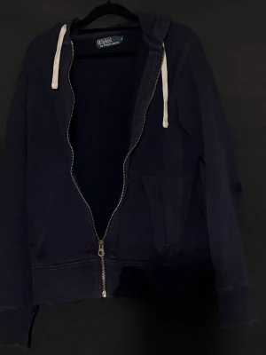 Polo Ralph Lauren - Marinblå Polo zip hoodie. Tröjan har ett smått  hål vid början av ärmen samt ett till litet vid armbågen, men något som inte syns så noga. I hel het är det en väldigt stilren och riktigt skön hoodie.