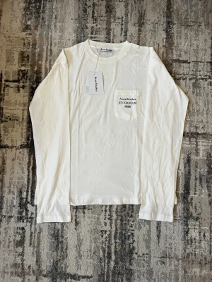 Vit långärmad t-shirt Acne Studios - Vit långärmad t-shirt från Acne Studios med bröstficka och svart text 'Acne Studios STOCKHOLM 1996' broderad på fickan. Klassisk rund halsringning och mjukt material, perfekt för en clean och stilren look.