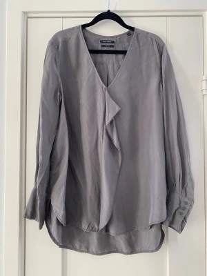 Grå blus med volang från Marc O'Polo i silke - Stilren grå blus från Marc O'Polo med v-ringning och dekorativ volang framtill. Blusen har långa ärmar med breda manschetter och knappar. Tillverkad i ett mjukt, lätt material som ger ett avslappnat men ändå elegant intryck. Sparsamt använd och i fint begagnat skick   Mått Över bröstet: ca 57 cm Längd från axeln och ner: ca 66 cm  100% silke  Kommer från ett djur och rökfritt hem