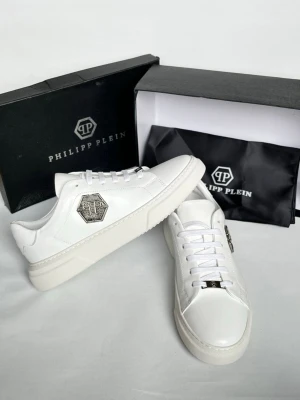 Vita Philipp Plein sneakers i läder - Stilrena vita sneakers från Philipp Plein i slätt läder med metallisk logga på sidan och detaljer på snörningen. Klassisk låg modell med tjock vit sula och rundad tå. Perfekta för dig som gillar exklusiv och clean streetstyle.