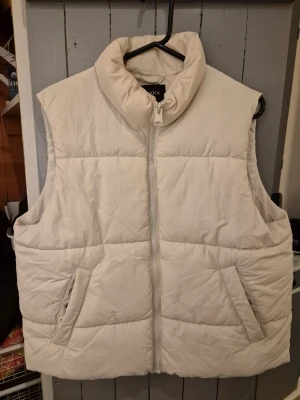 Beige quiltad pufferväst från Lindex - Snygg vit dunväst från Index med hög krage och dragkedja framtill. Västen är quiltad och har två praktiska fickor på sidorna. Perfekt att slänga över en hoodie för extra värme under kyliga dagar.