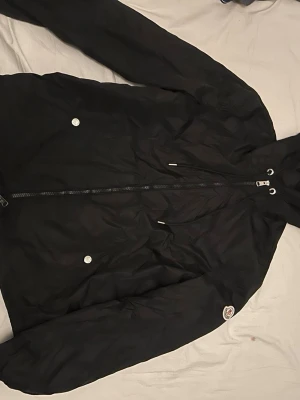  Moncler Grimpeurs Windbreaker - Snygg svart vindjacka från Moncler med huva och dragkedja framtill. Jackan har två fickor med tryckknappar och Moncler-logga på ärmen. Tillverkad i lätt och slitstarkt material som passar perfekt för blåsiga dagar.