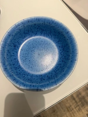 Blå skål från GN Sweden Granola - Säljer en rund skål i keramik från GN Sweden Granola. Skålen är blå med ett stänkmönster i mörkare blå nyanser och har en matt finish. Perfekt till frukost eller snacks. Märkt undertill med logga och text.