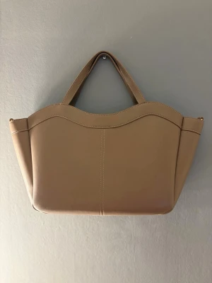 Beige, brun handväska i konstläder - Väskan har en mjuk, slät yta i konstläder och minimalistisk design utan synliga loggor. Perfekt för dig som gillar enkel och modern stil. En snygg brun/beige väska i fint skick 🤎✨   Perfekt storlek för både vardag och utflykter – rymmer allt du behöver!  👜 Stil: Tidlös och lätt att matcha   👜 Perfekt till: Skola, stan eller resor    Väskan är både praktisk och stilren- passar nästan alla outfits. 📦 Kan skickas eller mötas upp 💬 Hör av dig vid frågor eller intressen! 