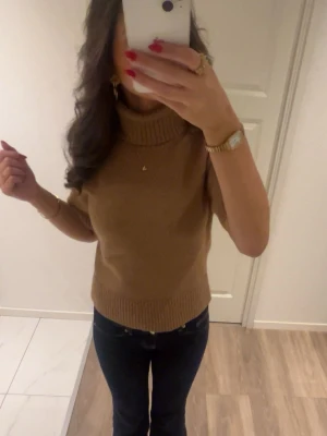 Beige stickad polotröja - Säljer en beige stickad polotröja med korta ärmar och ribbade detaljer vid krage, ärmslut och nederkant. Tröjan har en avslappnad passform och är perfekt att styla med jeans eller kjol. Materialet ser mjukt och bekvämt ut.