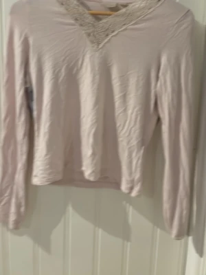 Ljusrosa långärmad topp med spets - Söt ljusbeige långärmad topp med v-ringning dekorerad med spetsdetalj. Perfekt för dig som gillar en feminin och enkel stil. Mjuk och skön känsla, passar till många olika outfits.