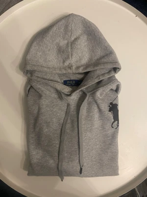 Grå hoodie från Polo Ralph Lauren - M passar S. Snygg grå hoodie från Polo Ralph Lauren med stor svart logga på bröstet och siffran 3 på ärmen. Klassisk huva med dragsko och ribbade muddar vid ärmar och nederkant. Perfekt för en avslappnad och trendig look. 