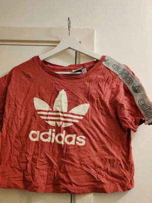 Croppad Adidas t-shirt vintage 38 40 - Säljer en röd croppad t-shirt från Adidas med stort vitt tryck av Adidas-loggan framtill och ett vitt band med Adidas-tryck på ena axeln. T-shirten har korta ärmar och är gjord i mjuk bomull. Perfekt för dig som gillar sportig och avslappnad stil.