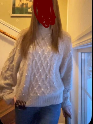 Beige stickad tröja med pärlor - Mysig beige stickad tröja med pärldetaljer och kabelstickat mönster framtill. Tröjan har rund hals, långa ärmar och ribbade muddar. Perfekt för kalla dagar och ger en söt, trendig look.