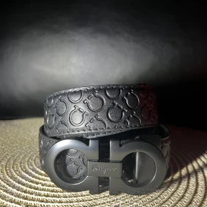 Bälte från Salvatore Ferragamo 105cm/42 - Snyggt svart bälte från Salvatore Ferragamo med präglat mönster och ikoniskt dubbel-Gancio-spänne. Tillverkat i skinn med en matt finish och stilren design som ger en lyxig känsla till outfiten.