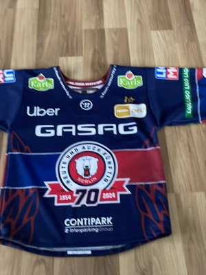 Eisbären Berlin hockeytröja blå - Säljer en officiell Eisbären Berlin hockeytröja i blått med röda och vita detaljer. Tröjan har tryckta sponsorer, klubbmärke och nummer 92 med namnet Noebels på ryggen. Materialet är polyester och modellen är loose fit, perfekt för hockeyfans. Det är en jubileums tröja från säsongen 2024/2025! Köpt i Berlin och nypris 1200kr!