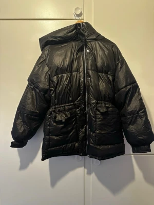 Svart pufferjacka från Bershka - Svart pufferjacka från Bershka med huva och dragkedja framtill. Jackan har två stora fickor med lock och justerbar midja. Perfekt för kalla vinterdagar och har en klassisk oversized look. Materialet är glansigt och ger en modern vibe.