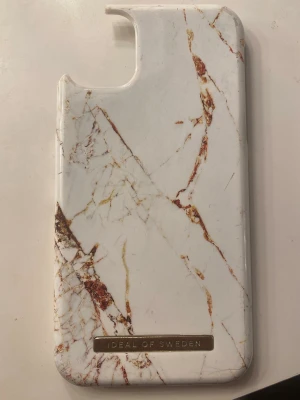 Mobilskal iDeal of Sweden - Snyggt mobilskal från iDeal of Sweden med vitt marmormönster och inslag av bruna och guldiga ådror. Skalet är gjort i hårdplast och har en elegant metallbricka med loggan längst ner. Perfekt för dig som vill ha en stilren och trendig look på mobilen. Passar till iphone 11🤍