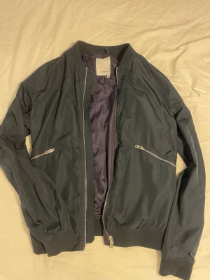 Mörkblå bomberjacka J.Lindeberg - Snygg mörkblå tunn bomberjacka från J.Lindeberg med silvriga dragkedjor och ribbade muddar. Storlek S