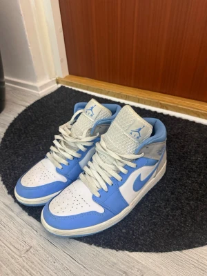 Nike Air Jordan 1 Mid UNC blå/vit - Säljer ett par Nike Air Jordan 1 Mid i färgen UNC blå och vit. Skorna har klassisk hög siluett, vit tåbox och blå detaljer samt swoosh. Ovandelen är i läder med mesh på plösen och Air Jordan-logga på sidan. Perfekta för dig som gillar streetwear och sneakerskultur.