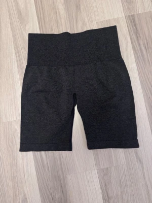 Mörkgrå höga cykelshorts - Mörkgrå höga cykelshorts med bred midjeresår och diskret logga på baksidan. Tillverkade i stretchigt material som passar perfekt för träning eller en aktiv vardag. Snygg och enkel design utan synliga sömmar framtill.