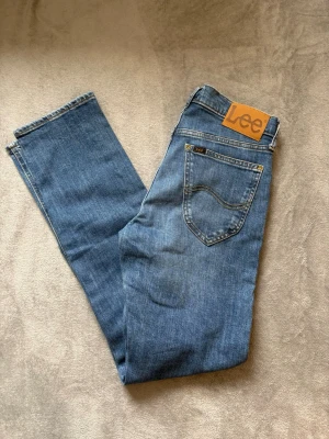 Herr blå jeans från Lee - Snygga ljusblå Lee-jeans med klassisk design. Rak passform och normal midja. Knäppning fram och Lee-logga på bakfickan. Väldigt bra skick i storlek W28/L32. Original pris är 1200kr