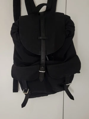 Svart ryggsäck med spännen - Stilren svart ryggsäck i tyg med detaljer i konstläder. Ryggsäcken har två framfickor med spännen och ett lock som stängs med rem och metallspänne. Justerbara axelband och praktiskt handtag upptill. Perfekt för dig som gillar enkel och klassisk design.