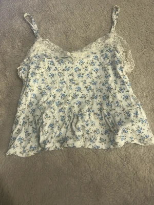 Blommig spetslinnetopp med volang - Söt linnetopp med blått blommönster på vit botten och spetsdetaljer vid urringningen. Toppen har smala axelband och volangkant nedtill för en romantisk vibe. Perfekt för varma dagar och sommarens alla äventyr. 