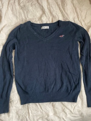 Mörkblå stickad tröja Hollister - Mörkblå stickad tröja från Hollister med V-ringning och lång ärm. Tröjan har ribbade muddar vid ärmslut och nederkant samt en liten röd logga broderad på bröstet. Perfekt för lager på lager och enkel att matcha med jeans. Skriv om ni har andra frågor!🥰