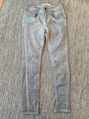 Grå jeans med blå detaljer Mos Mosh - Snygga grå jeans från Mos Mosh med raka ben och coola blå broderade detaljer vid fickorna fram. Klassisk femficksmodell med knapp och dragkedja. Perfekta för dig som vill ha en unik twist på dina vardagsjeans.Ett par gråblå byxor från Mos Mosh med dekorativa detaljer på fickorna fram och bak. Byxorna har en smal passform.  Nya, aldrig använda  Mått Midjan: ca 37 cm Längd från grenen och ner: ca 77 cm  97% bomull och 3% elastan  Kommer från ett djur och rökfritt hem