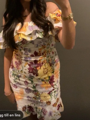 Blommig offshoulder klänning - Säljer en söt blommig offshoulder klänning med volangdetaljer upptill och nertill. Klänningen har en vit bas med färgglada blommor i gult, lila och grönt. Perfekt för sommarens alla tillfällen!