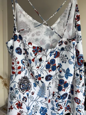 Blommig linnetopp med tunna band - Säljer en vit linnetopp med tunna axelband och blommigt mönster i blått, rött. Toppen har korsade band i ryggen. Perfekt för varma dagar och enkel att styla till jeans eller kjol.