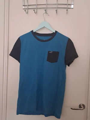 Blå t-shirt - Blå t-shirt från Hollister med kontrasterande mörkgrå ärmar och bröstficka. T-shirten har rund halsringning och en liten Hollister-logga på fickan. Se bilder för skick 🩷 Uppskattad storlek M. 