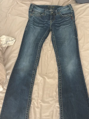 Blå bootcut jeans från Silver - Snygga blå bootcut jeans från Silver med slitningar och kontrastsömmar. Klassisk femficksmodell och låg midja. Jeansen har detaljerade sömmar och logga på bakfickan. W28 L33