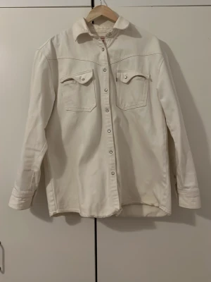 Vit overshirt med tryckknappar (L) - Stilren vit Jeansjacka i kraftigt bomullstyg med klassisk krage och snygga tryckknappar framtill vilket gör det lättare att knäppa jackan. Två bröstfickor med lock och knapp, samt långa ärmar med manschett. Perfekt lager-på-lager-plagg för en avslappnad look. Märket är Levi’s och Jeansjackan kommer hålla i många år. Kom ihåg att när du alltid köper från mig kan vi diskutera priset. Hör gärna av dig! 