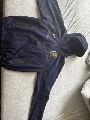 Mörkblå AIK vindjacka från Nike - Mörkblå vindjacka med huva från Nike, prydd med AIK:s klubbmärke och gul Nike-logga på bröstet. Jackan har två snedställda dragkedjefickor och reflekterande detaljer. Perfekt för dig som vill visa ditt AIK-supporterhjärta.