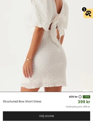 Vit kort klänning med rosett - Trendig vit kort klänning med struktur, puffärmar och stor rosett i ryggen. Klänningen har en öppen rygg och är figurnära med en mjuk, texturerad yta. Perfekt för dig som vill sticka ut med en söt och modern look.