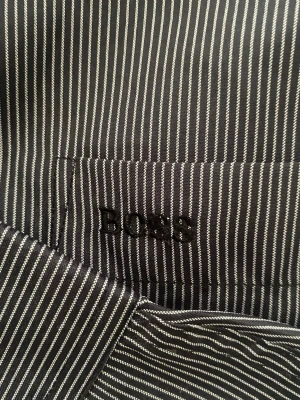 Svartvit randig skjorta från BOSS - Snygg svart skjorta från BOSS med tunna vita ränder och klassisk krage. Skjortan har långa ärmar, bröstficka med diskret BOSS-brodyr och knäppning framtill. Perfekt för dig som gillar stilrena detaljer och tidlös design.