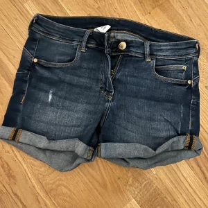 Mörkblå jeansshorts med uppvik - Snygga mörkblå jeansshorts med uppvikta benslut och klassisk femficksdesign. Shortsen har gylf med dragkedja och knapp samt kontrastsömmar. Använt fint skick. Stl:34/36. Upplever dem som rätt små i modellen. Elastiska 