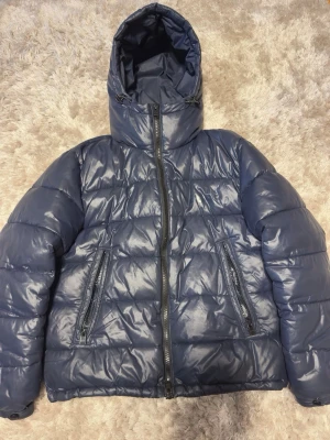 Mörkblå pufferjacka från BOSS - Snygg mörkblå pufferjacka från BOSS med huva och dragkedja framtill. Jackan har två framfickor med dragkedja och quiltad design. Materialet är glansigt och ger en modern look, perfekt för kalla vinterdagar.