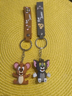 Tom & Jerry nyckelringar i gummi - Oanvända. Två färgglada nyckelringar med Tom & Jerry-tema. Ena är brun med Jerry och den andra grå med Tom, båda i mjukt gummi och med tryckt text och figurer. Perfekt accessoar för dig som gillar klassiska tecknade serier.