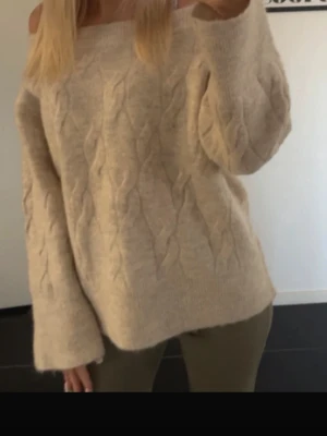 Beige off shoulder stickad tröja - Mysig beige stickad tröja med kabelstickat mönster och off shoulder-design. Tröjan har långa ärmar och en avslappnad passform, perfekt för lager på lager. Materialet ser ut att vara mjukt och fluffigt, vilket ger en skön känsla.