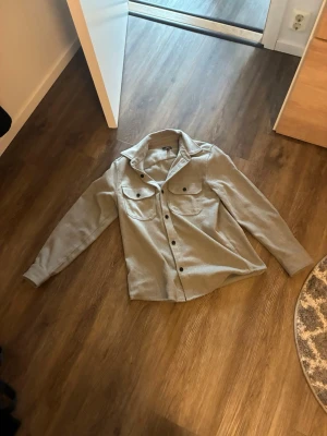 Grå overshirt från H&M - Stilren grå overshirt från H&M i regular fit. Skjortjackan har två bröstfickor med lock och knappar framtill. Perfekt lager-på-lager-plagg med klassisk krage och långa ärmar. Materialet känns mjukt och passar till många olika outfits.