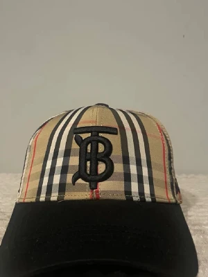 Burberry keps - Snygg keps från Burberry med det ikoniska beige, svarta, vita och röda rutmönstret. Framtill finns en stor broderad TB-logga i svart. Skärmen är svart och kepsen har justerbart spänne bak. Tillverkad i bomull för en bekväm känsla.