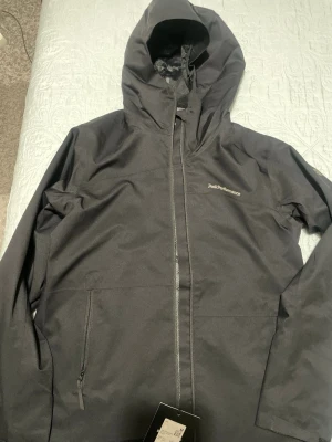 Svart vindjacka från Peak Performance - Snygg svart vindjacka från Peak Performance med huva och dragkedja framtill. Jackan har två sidofickor med dragkedja och diskret logga på bröstet. Tillverkad i ett slitstarkt, vattenavvisande material som passar perfekt för blåsiga dagar. Pris går o diskutera