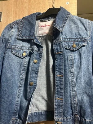 Blå jeansjacka från John Baner - Klassisk blå jeansjacka från John Baner med snygga bronsfärgade knappar och två bröstfickor med lock. Jackan har en rak passform, markerad krage och tydliga sömmar som ger en tidlös look. Perfekt att slänga över en hoodie eller t-shirt.