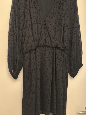 Svart omlottklänning med mönster - Svart omlottklänning med subtilt blommönster och v-ringning. Klänningen har långa, lösa ärmar och ett mjukt, flytande tyg som ger en avslappnad men elegant look. Perfekt för dig som gillar stilrena detaljer och en bekväm passform.