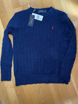Marinblå kabelstickad tröja Polo  - Kabelstickad marinblå tröja från Polo med rund halsringning och den klassiska röda logotypen broderad på bröstet. Tröjan har långa ärmar och ribbade muddar vid ärmslut och nederkant. Perfekt för en stilren och avslappnad look.