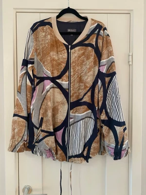 Mönstrad bomberjacka med dragkedja - Unik bomberjacka från Ulla Popken med abstrakt mönster i beige, marinblått, lila och vitt. Jackan har dragkedja framtill, snörning i nederkant och långärmad design. Perfekt för dig som vill sticka ut med en färgstark och bekväm jacka.Ny beige mönstrad glansig jacka i strl 50/52 fr Ulla Popken   Ny, aldrig använd  Mått Över bröstet: ca 71 cm Längd från axeln och ner: ca 77 cm  Viskos, bomull, elastan och polyester   Kommer från ett djur och rökfritt hem