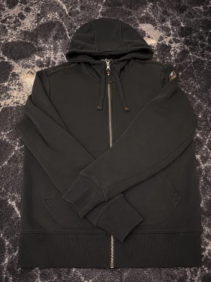 Svart hoodie från Parajumpers - Svart hoodie med dragkedja från Parajumpers. Hoodien har huva med dragsko, ribbade muddar och fickor framtill. Detaljerad med ett broderat Parajumpers-märke på ena ärmen och orange sömmar vid axlarna. Perfekt för en avslappnad stil. Priset är förhandlingsbart.