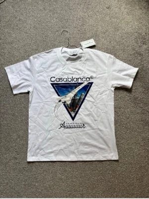 Vit Casablanca t-shirt med tryck - Vit t-shirt från Casablanca med stort grafiskt tryck framtill i blått och svart, samt texten 'Casablanca' och 'Aiiiiiiir'. Klassisk passform och rund hals. Perfekt för dig som gillar streetwear och snygga prints. S men sitter som L