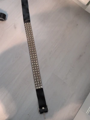 Svart nitar bälte med silverdetaljer - Säljer ett svart bälte med silvriga fyrkantiga nitar längs hela mittenpartiet. Bältet har ett klassiskt spänne i silver och är gjort i ett syntetmaterial. Perfekt för att ge din outfit en edgy vibe.