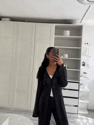 Svart glittrig lång cardigan - Snygg svart lång cardigan med glittriga detaljer. Jackan är tunn och har en öppen front med en knapp upptill. Materialet är genomskinligt och ger en trendig look, perfekt att slänga över en topp för extra stilpoäng. Långa ärmar och rak passform.
