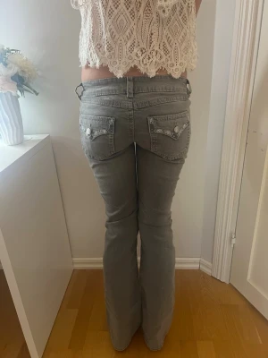 Grå bootcut jeans med detaljer - Säljer ett par grå bootcut jeans med snygga fickdetaljer och knappar bak. Jeansen har låg midja och klassisk femficksmodell. Materialet är denim med lite stretch för skön passform. Perfekta till en trendig vardagslook. Helt nya utan prislapp!